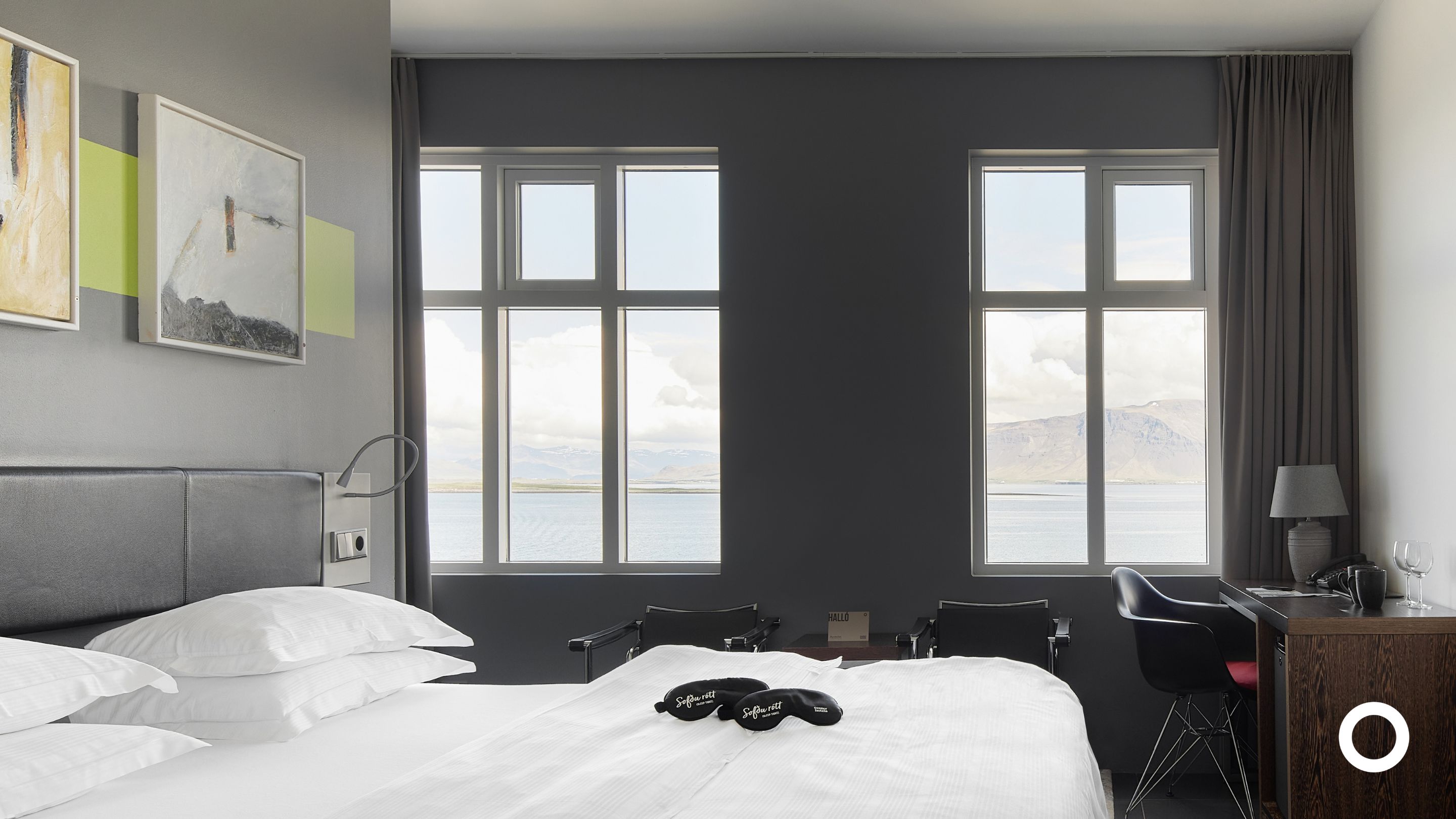 Ein Hotelzimmer mit Bett, zwei Stühlen, einem Schreibtisch und großen Fenstern mit Blick auf Berge und Wasser.