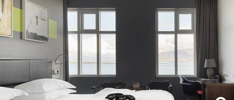 Ein Hotelzimmer mit Bett, zwei Stühlen, einem Schreibtisch und großen Fenstern mit Blick auf Berge und Wasser.