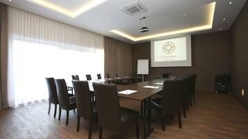 Et mødelokale med et langt bord, stole, et projektorskærm der viser 'Columbus Hotel' og en whiteboardtavle.