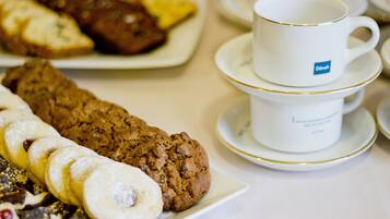 A table with a plate of pastries, a large cookie, and a stack of white cups with gold rims.