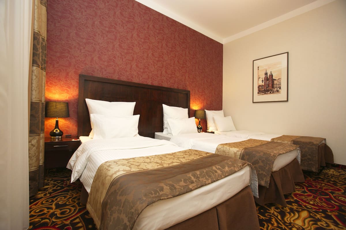 A hotel room with two beds, a red patterned wall, a framed cityscape picture, and a carpeted floor.