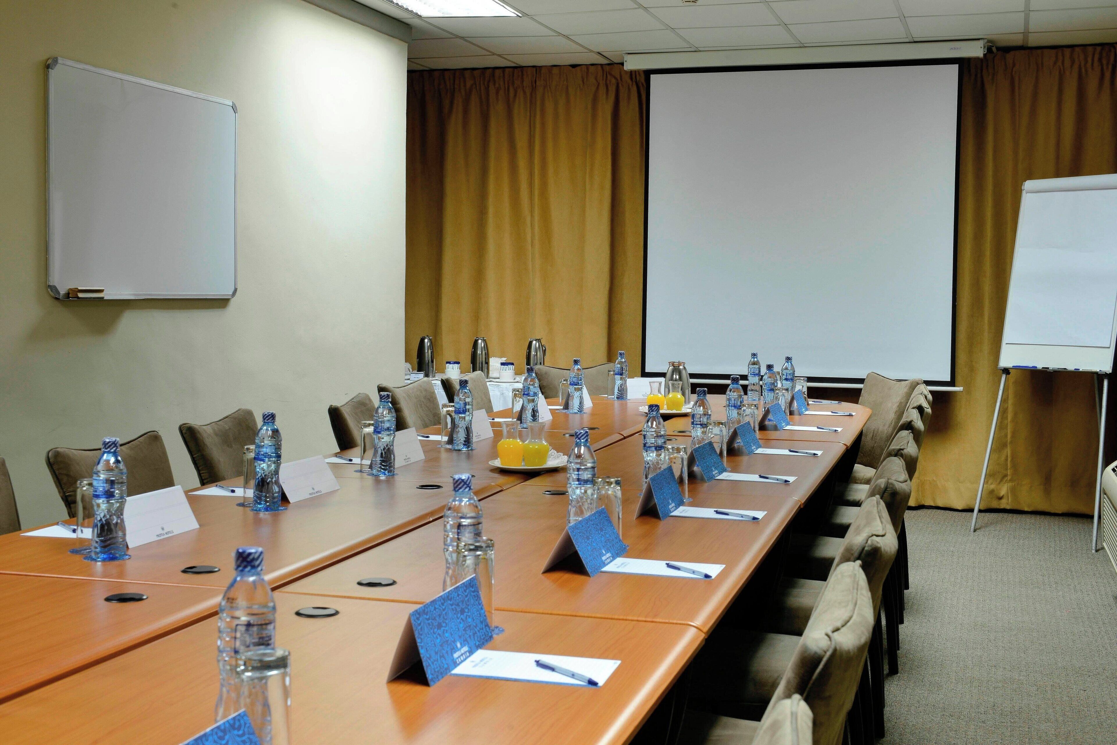 A conference room with a long table, chairs, nameplates, water bottles, and a projector screen.