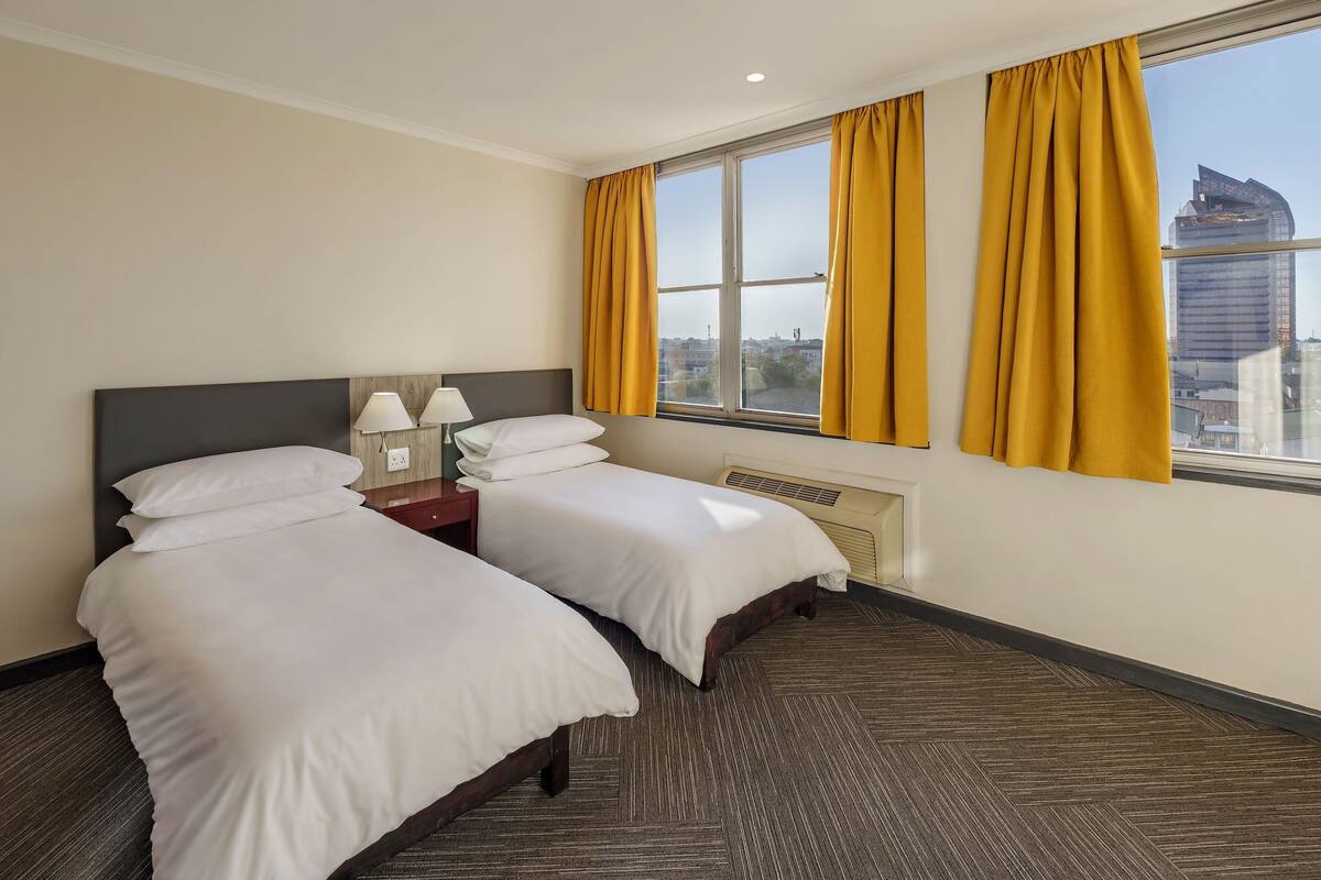 Une chambre d’hôtel avec deux lits, une fenêtre avec des rideaux jaunes, une lampe et une vue sur le paysage urbain.