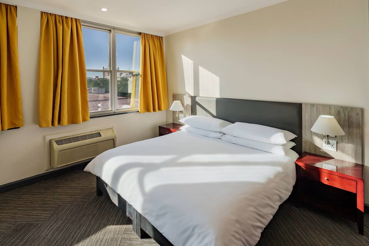 Une chambre d’hôtel avec un grand lit, deux tables de chevet avec des lampes, une fenêtre avec des rideaux jaunes et un radiateur.