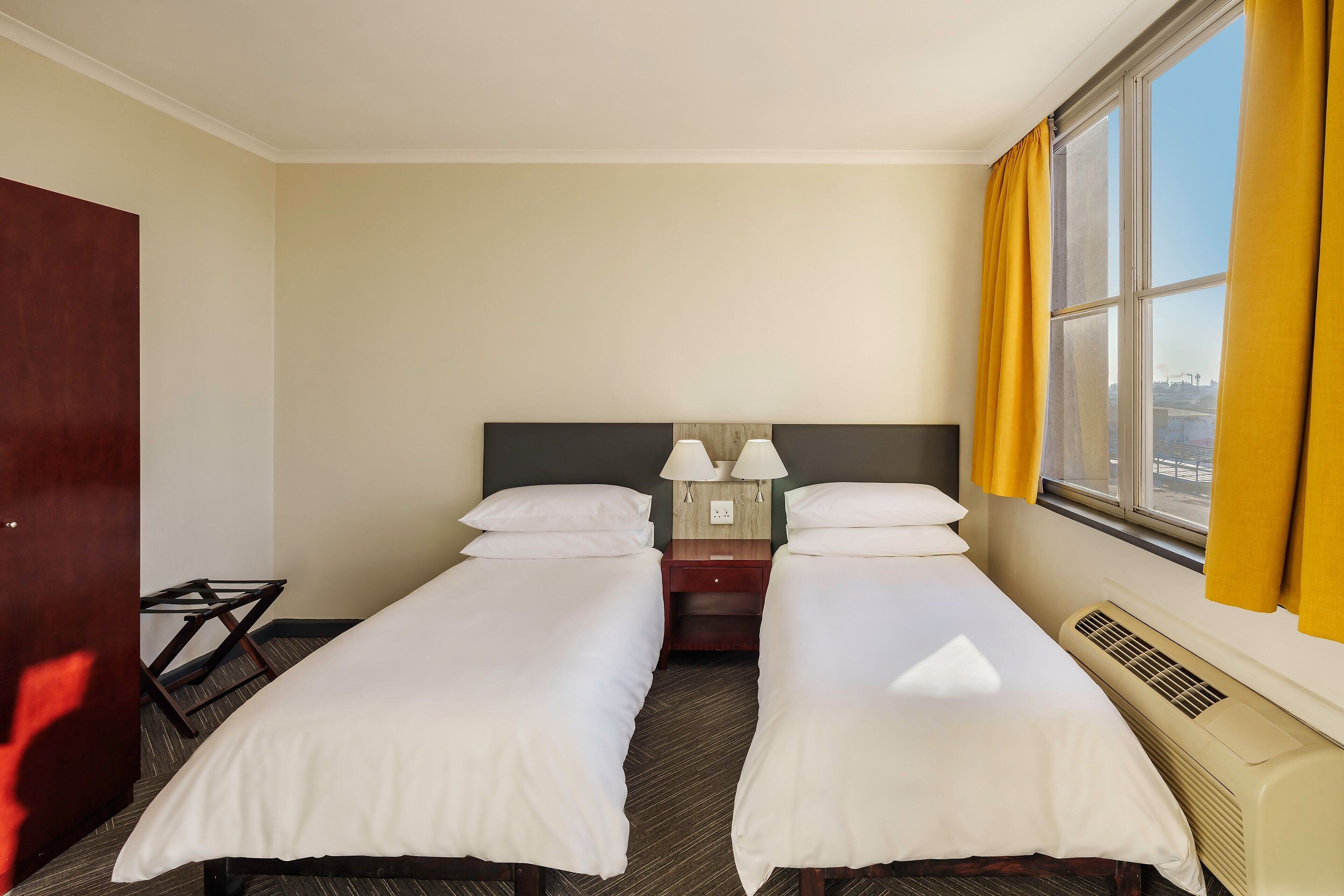 A hotel room with two single beds, a nightstand with lamps, a heater, and a window with yellow curtains.