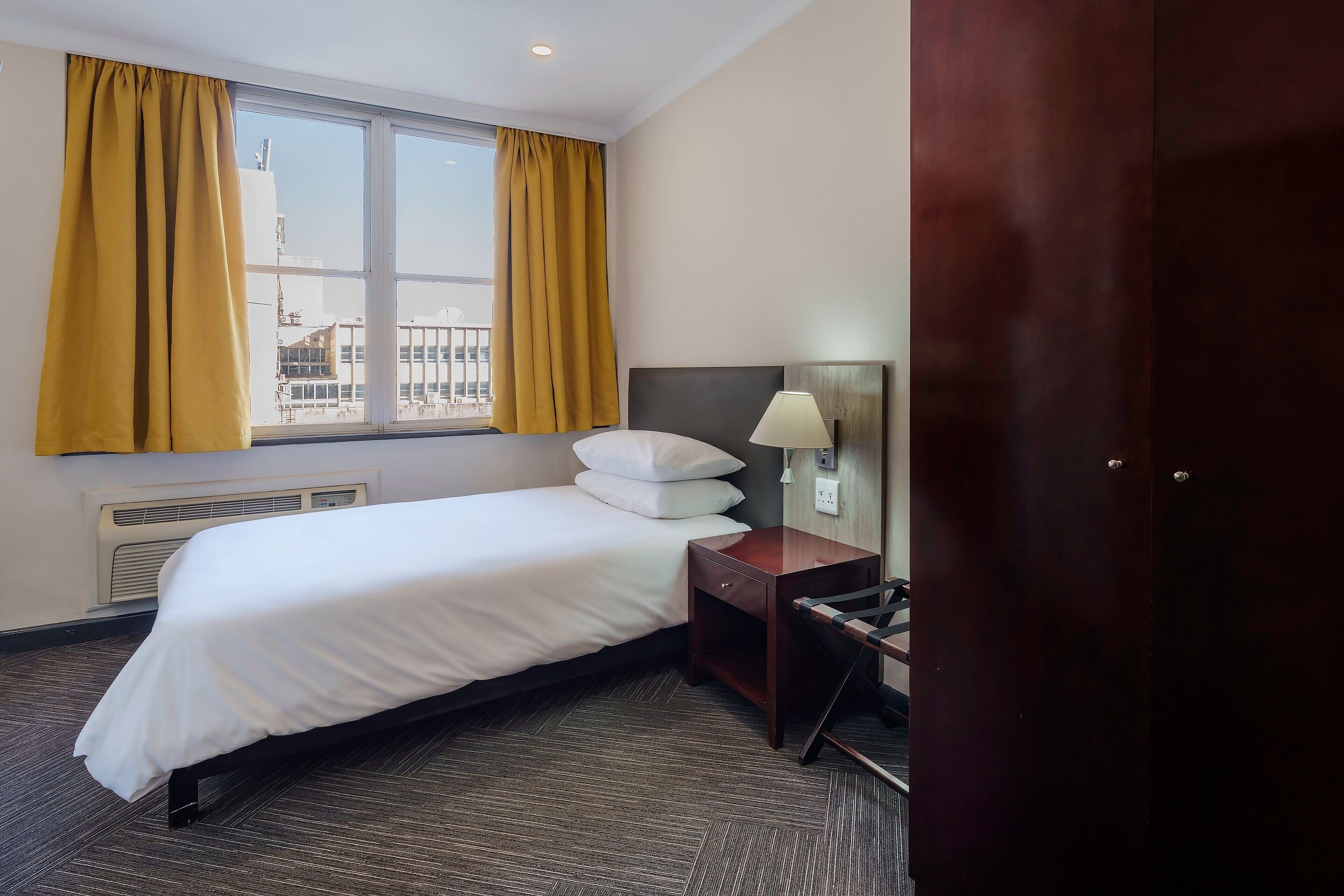Habitación de hotel con cama, mesita de noche con lámpara, ventana con cortinas y vista a edificios.