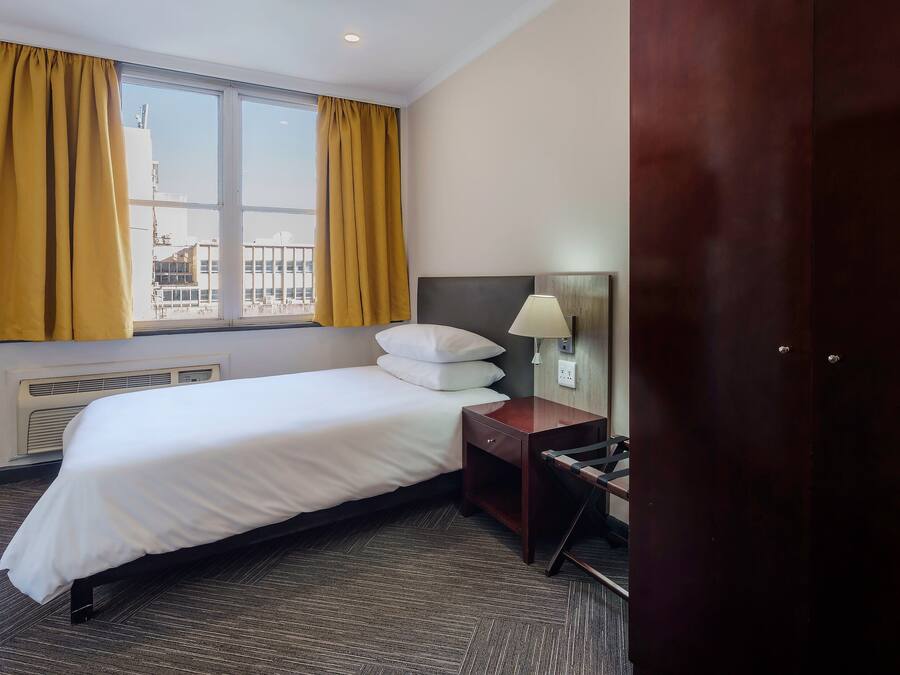Habitación de hotel con cama, mesita de noche con lámpara, ventana con cortinas y vista a edificios.