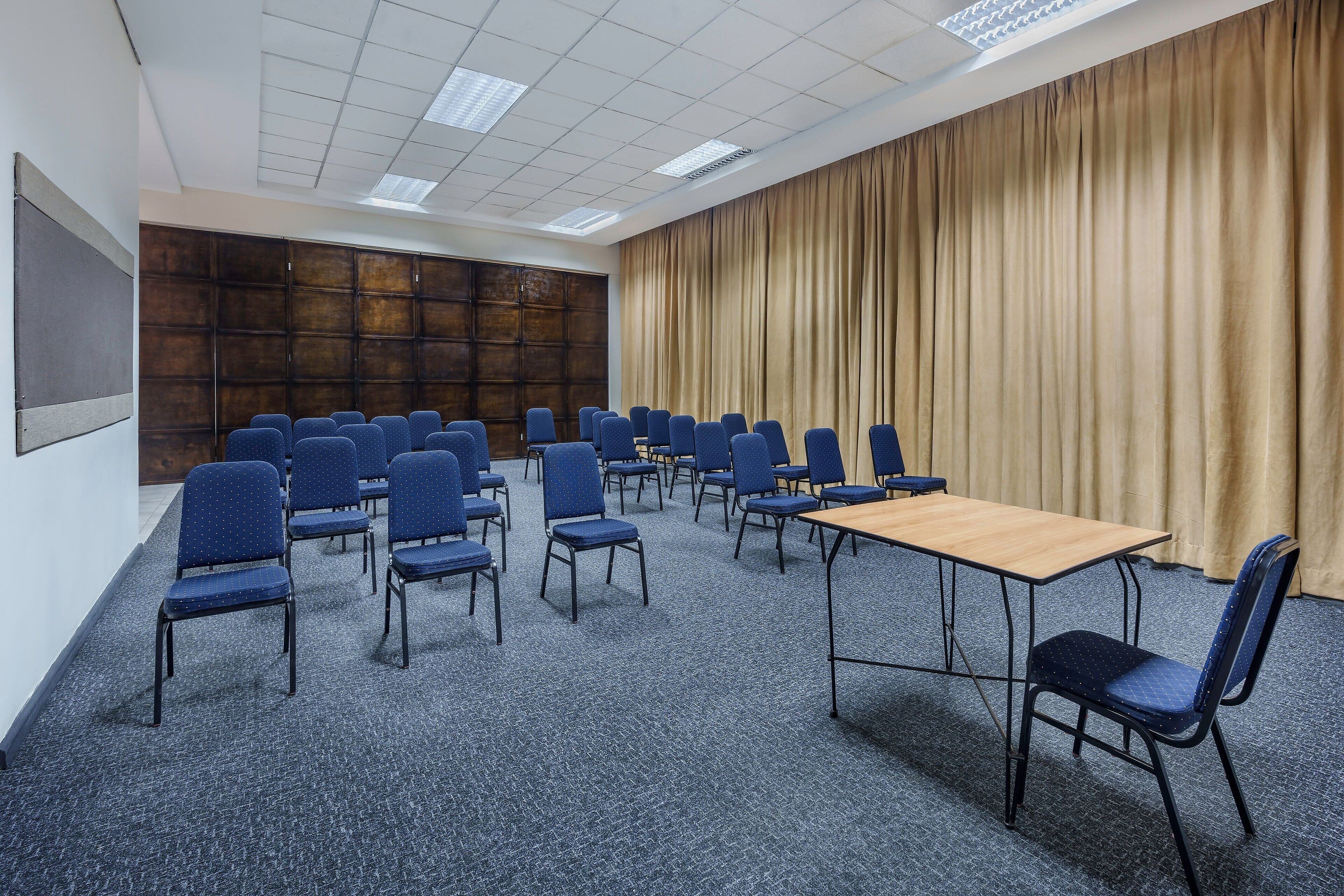 A conference room with rows of blue chairs, a wooden table, and a large curtain backdrop.