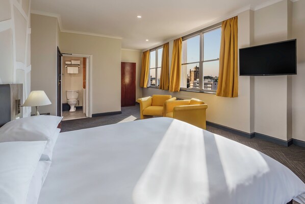 Habitación de hotel con una cama grande, televisor montado en la pared, ventana con cortinas y vistas a la ciudad.