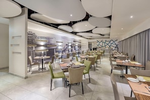 Un comedor moderno con varias mesas y sillas, una zona de bar central, y una pared con un mural de un paisaje urbano.
