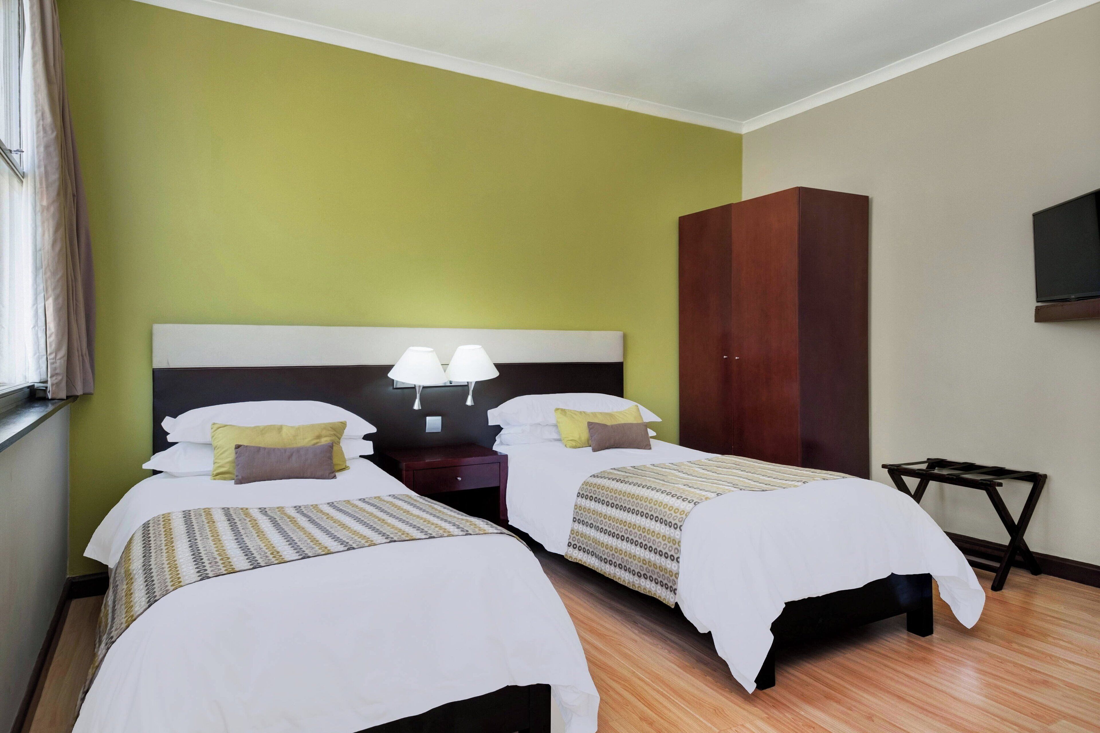 A hotel room with two beds, a nightstand with lamps, a wardrobe, and a TV mounted on the wall.