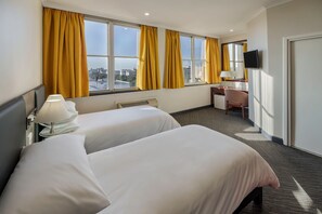 Une chambre d’hôtel avec deux lits, un bureau, une chaise, une télévision et de grandes fenêtres avec des rideaux.