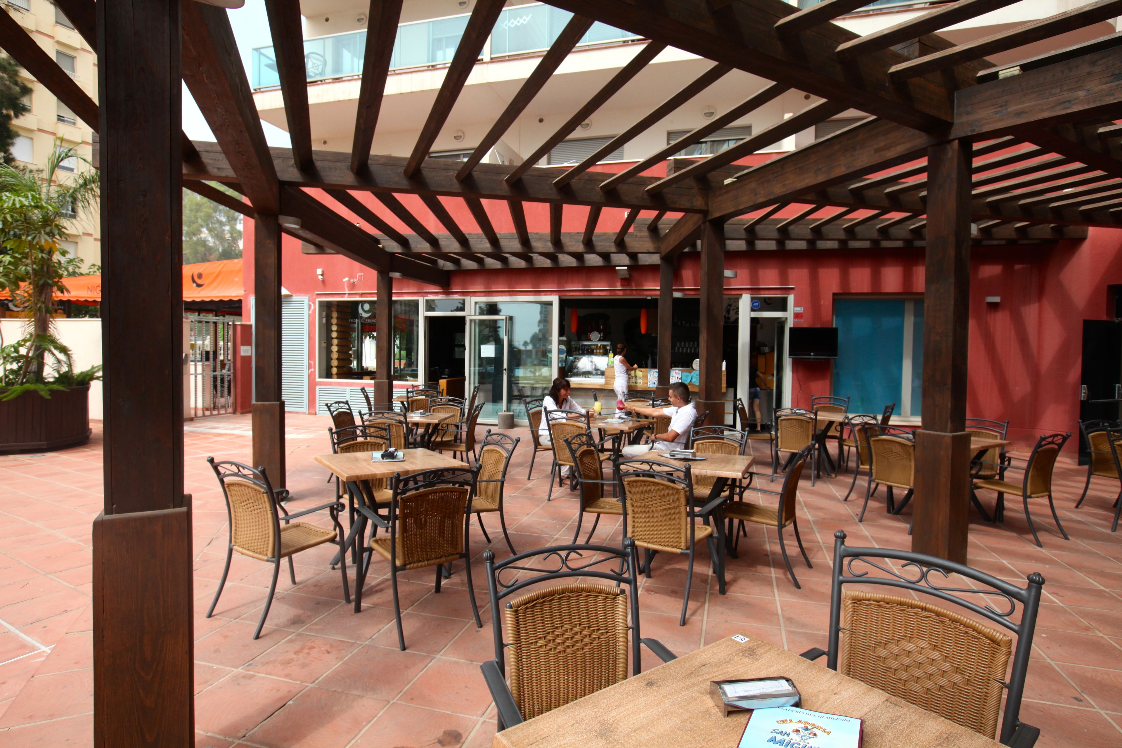 An outdoor café with wooden tables and wicker chairs, a covered area with a red wall, and a view of the street in the background.