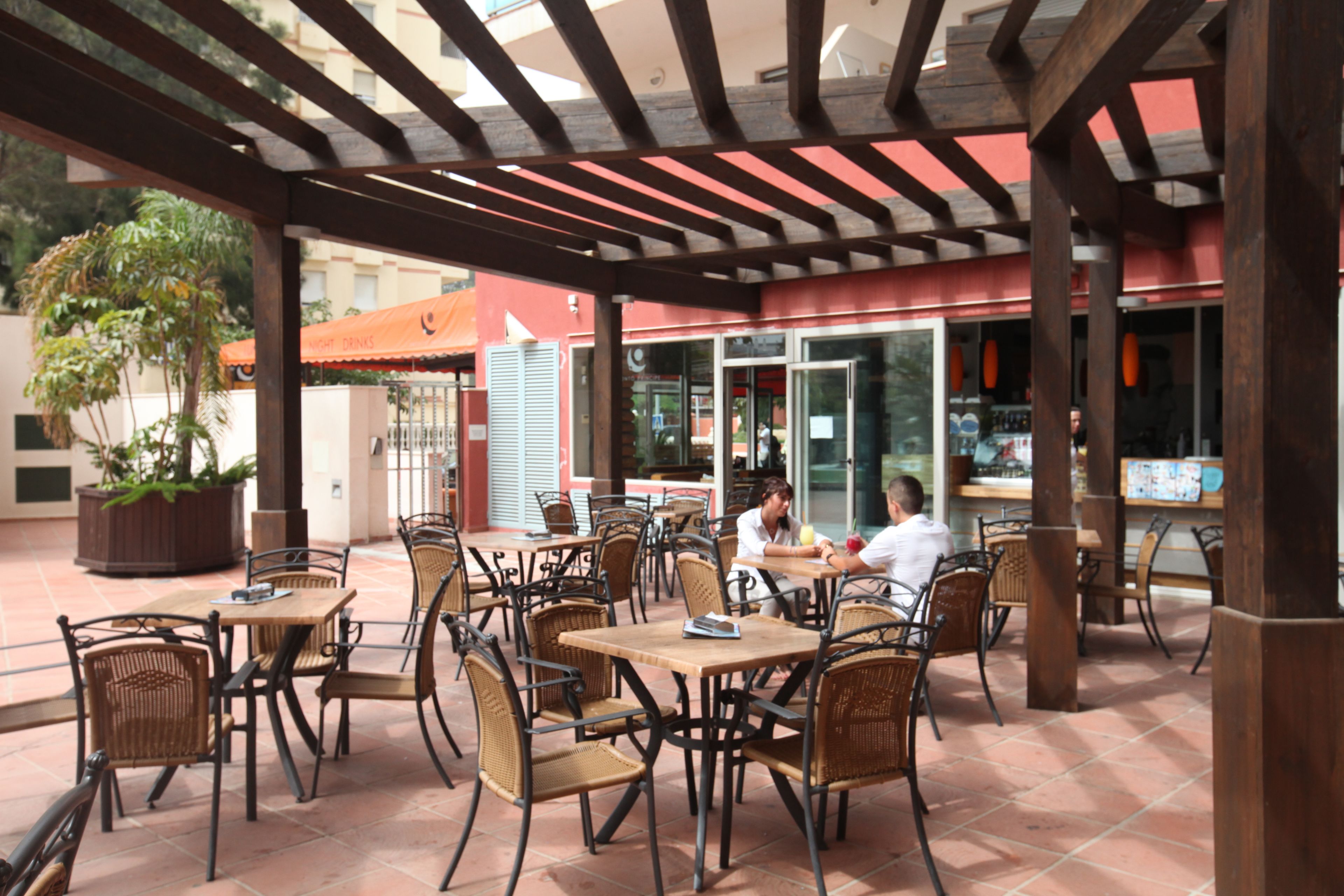 Un café en plein air avec des tables en bois et des chaises en osier, un sol carrelé et un espace abrité doté d’un plafond en bois.