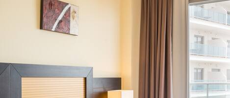 A hotel room with two beds, a headboard, a bedside table with a lamp, a window with curtains, and a painting on the wall.