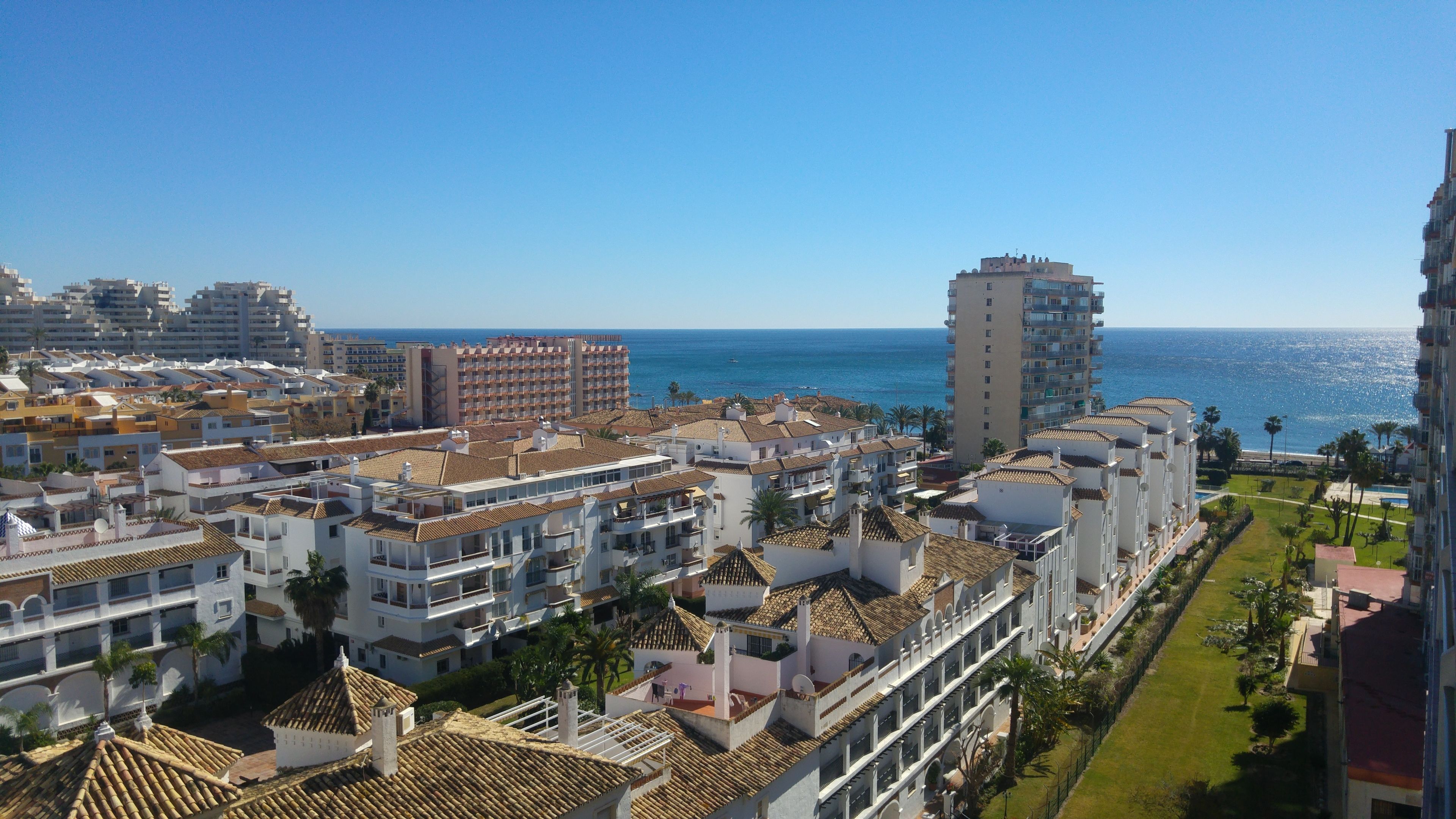 A coastal residential area with white buildings, some with red-tiled roofs, and a tall building with balconies overlooking the sea.