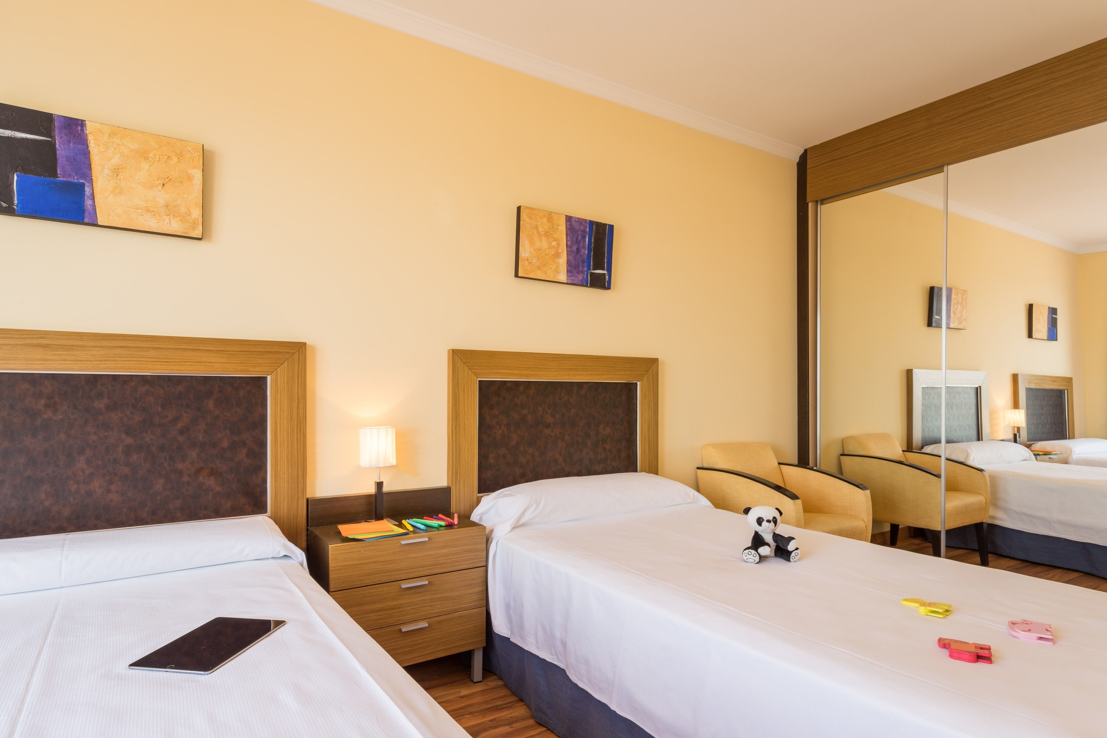 Una habitación de hotel con dos camas, una mesita de noche, una lámpara, una silla y obras de arte en las paredes.