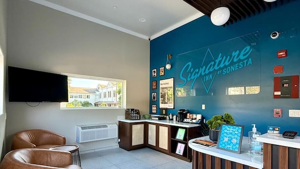 A reception area with a blue wall, wooden slats, a reception desk, a flat-screen TV, and a window with a view of a building. - Signature Inn Berkeley (Berkeley)
