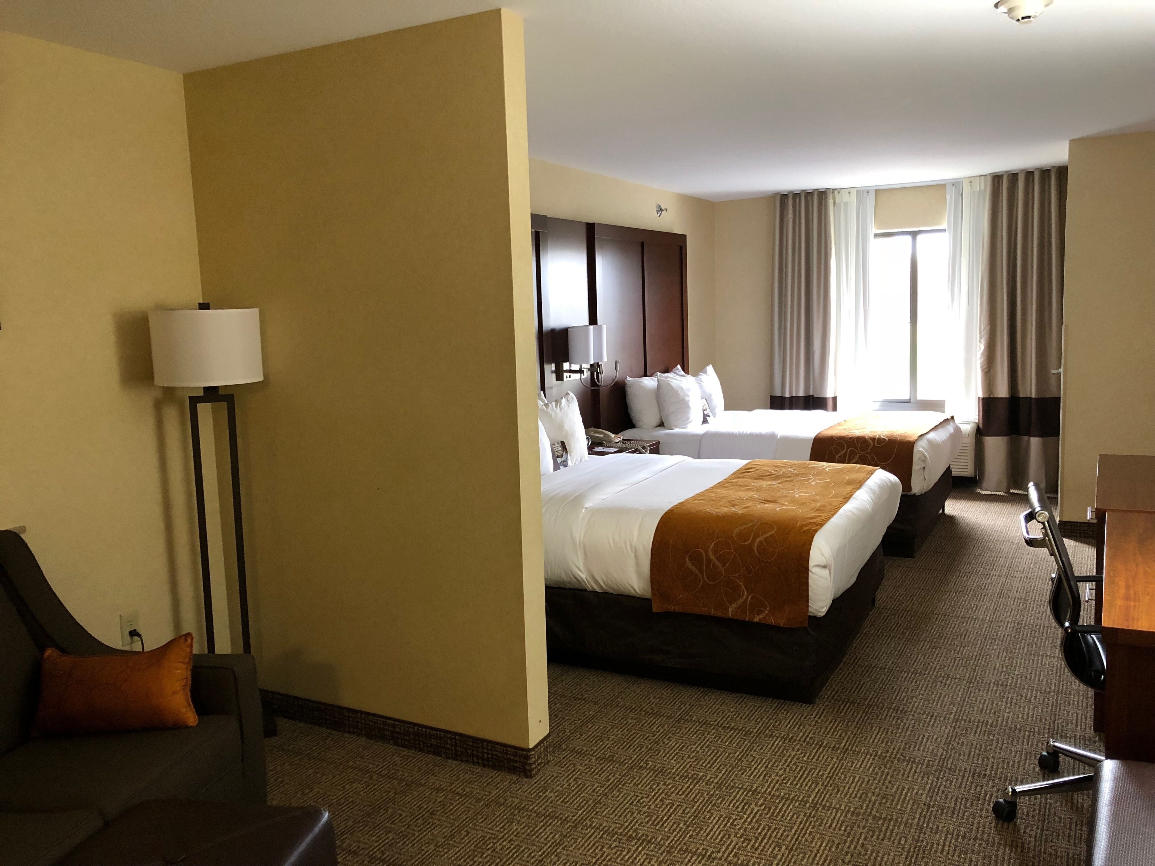 A hotel room with two beds, a desk, a chair, a lamp, and a view of the outdoors.