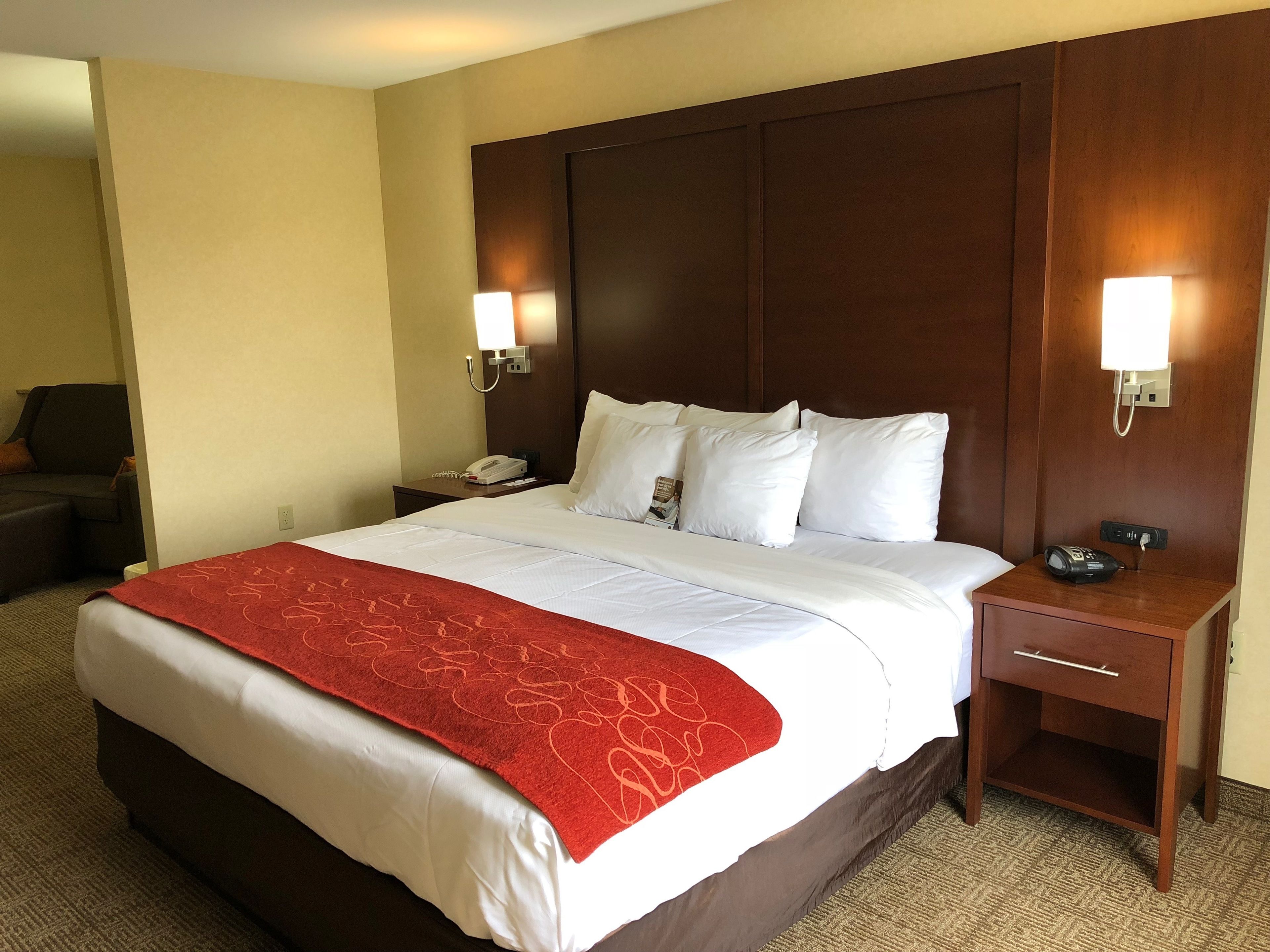 A hotel room with a large bed, a red throw blanket, a wooden headboard, two bedside lamps, a nightstand with a phone, and a small drawer.