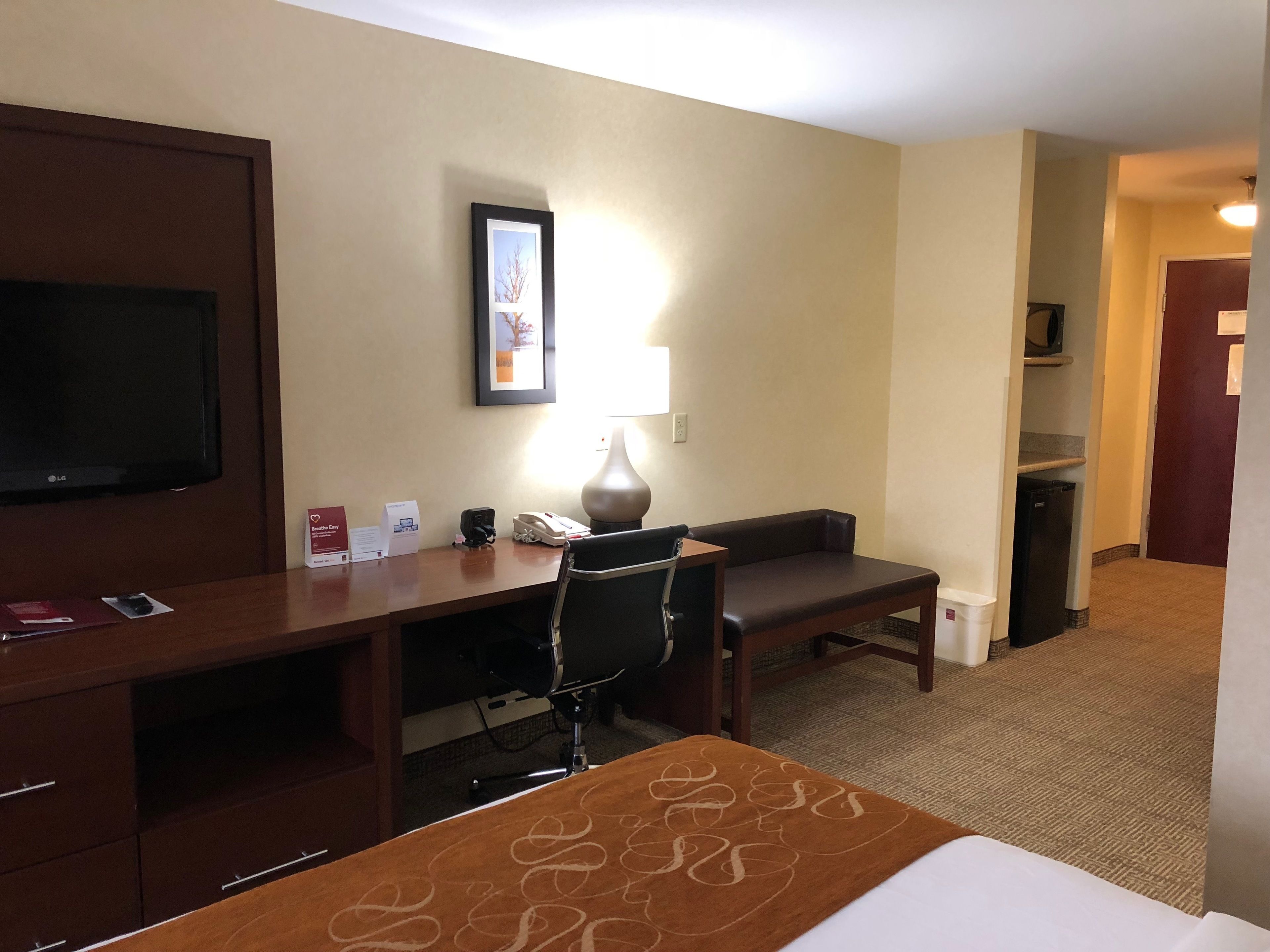 A hotel room with a bed, a desk with a chair, a television, a lamp, and a framed picture on the wall.