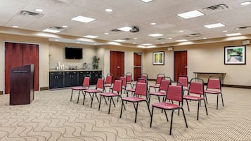 Une salle de conférence avec un tapis à motifs, des rangées de chaises rouges, un pupitre, un téléviseur et un tableau encadré accroché au mur.