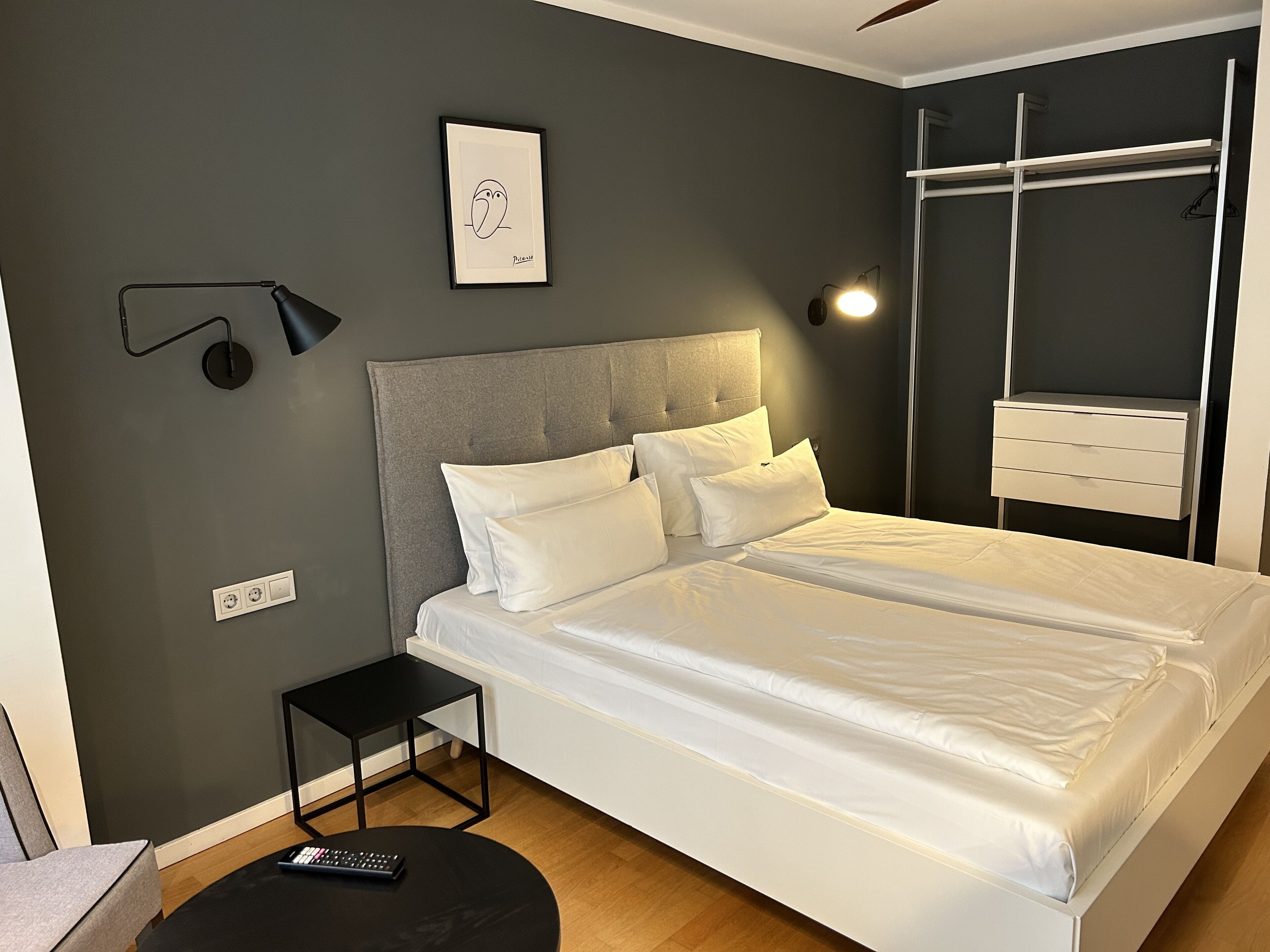 A modern bedroom with a large bed, a small side table, a wall-mounted light, and a framed picture.