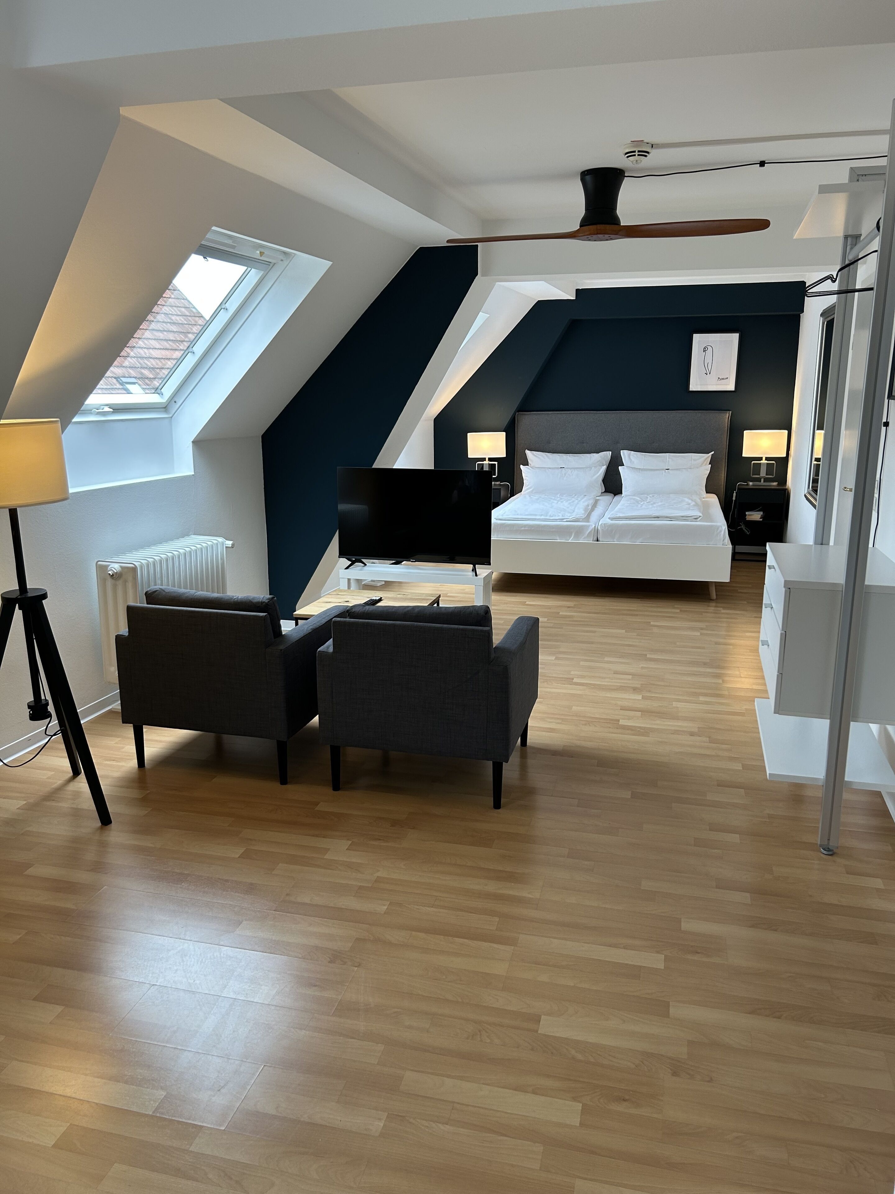 A modern hotel room with a loft-style ceiling, a large bed, a desk, and a seating area with two chairs.