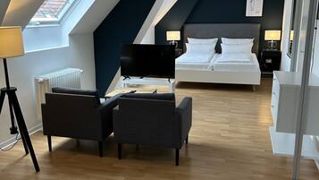 A modern hotel room with a loft-style ceiling, a large bed, a desk, and a seating area with two chairs.