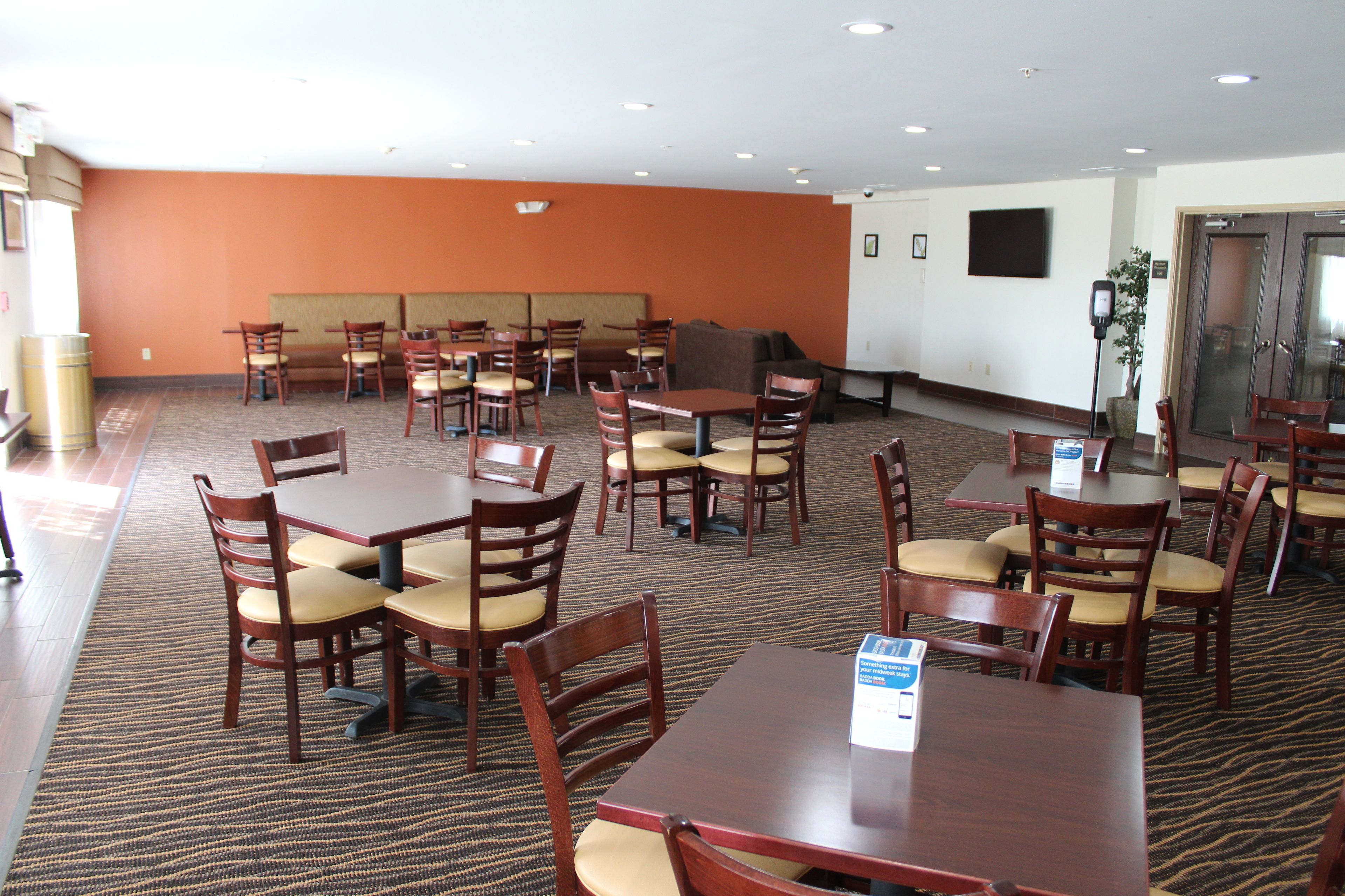A hotel dining area with multiple tables and chairs, a carpeted floor, and a television on the wall.