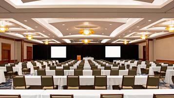 Une salle de conférence avec des rangées de tables et de chaises, une scène équipée d’un projecteur et d’un grand écran.