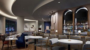 Un espacio moderno de bar con mesas redondas y taburetes, una barra con gran variedad de botellas y una zona de estar con sofás y sillones.