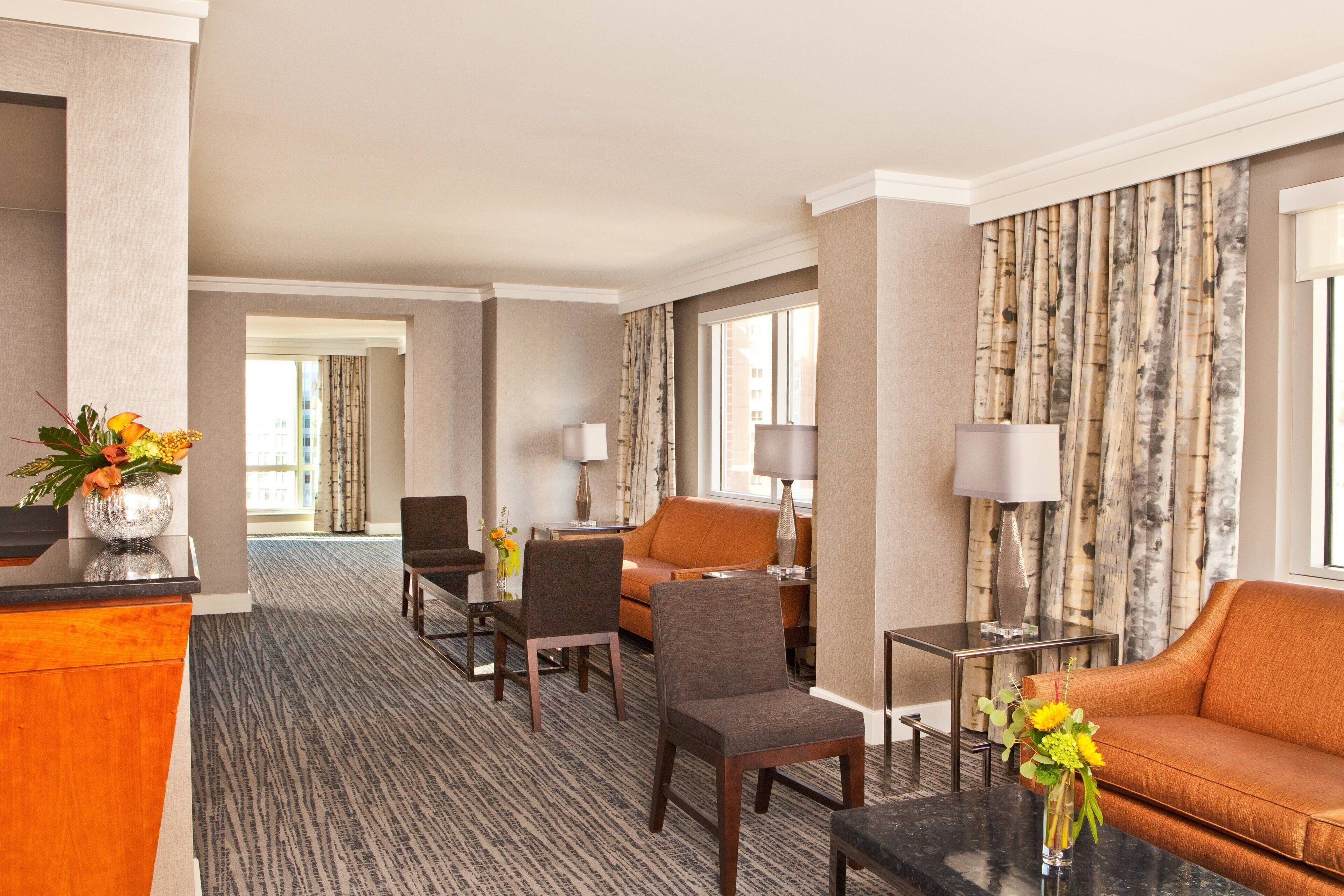 Habitación de hotel con mostrador de recepción, zona de estar con sofá y sillones, mesa de comedor y un amplio ventanal con cortinas.