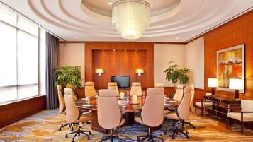 A conference room with a round table, beige chairs, a chandelier, a flat-screen TV, and a large painting on the wall.