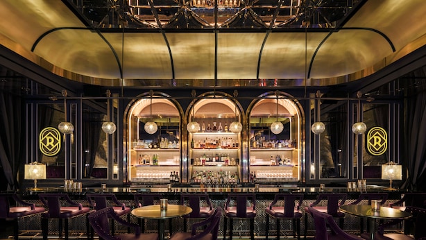 A bar with a high ceiling, ornate chandeliers, and a bar counter with a variety of bottles. There are multiple seating areas with tables and chairs.