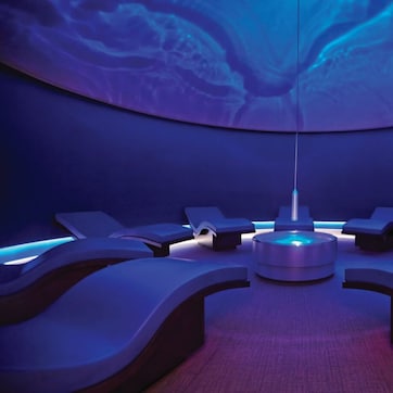 A modern spa room with a central circular light fixture, multiple reclining chairs, and a curved ceiling with a blue light design.