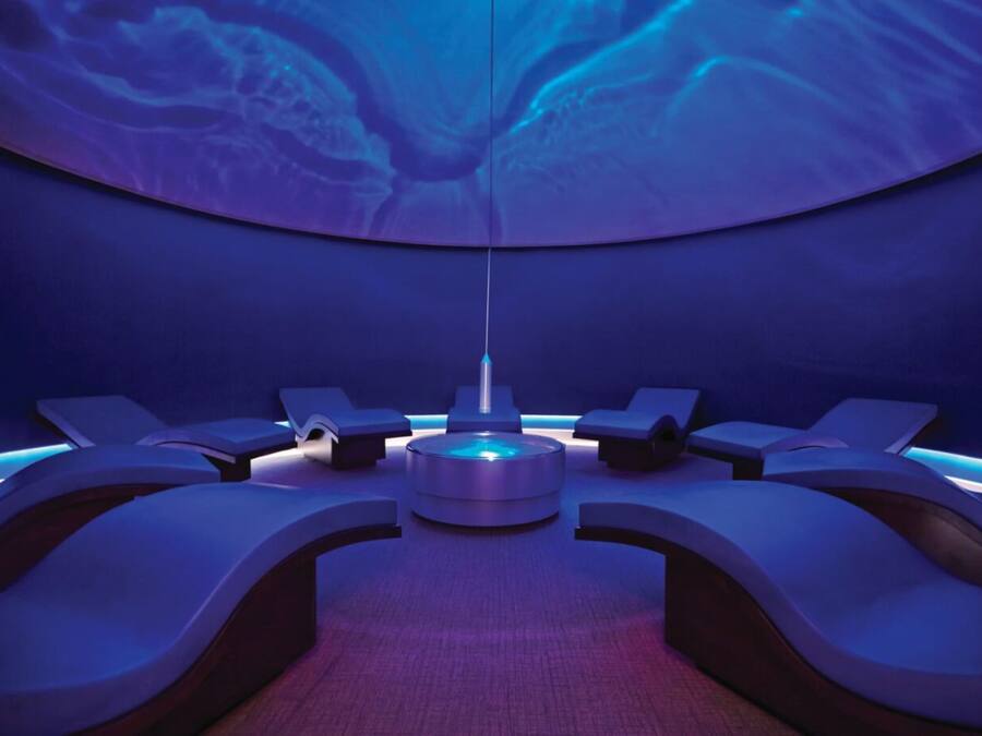A modern spa room with a central circular light fixture, multiple reclining chairs, and a curved ceiling with a blue light design.