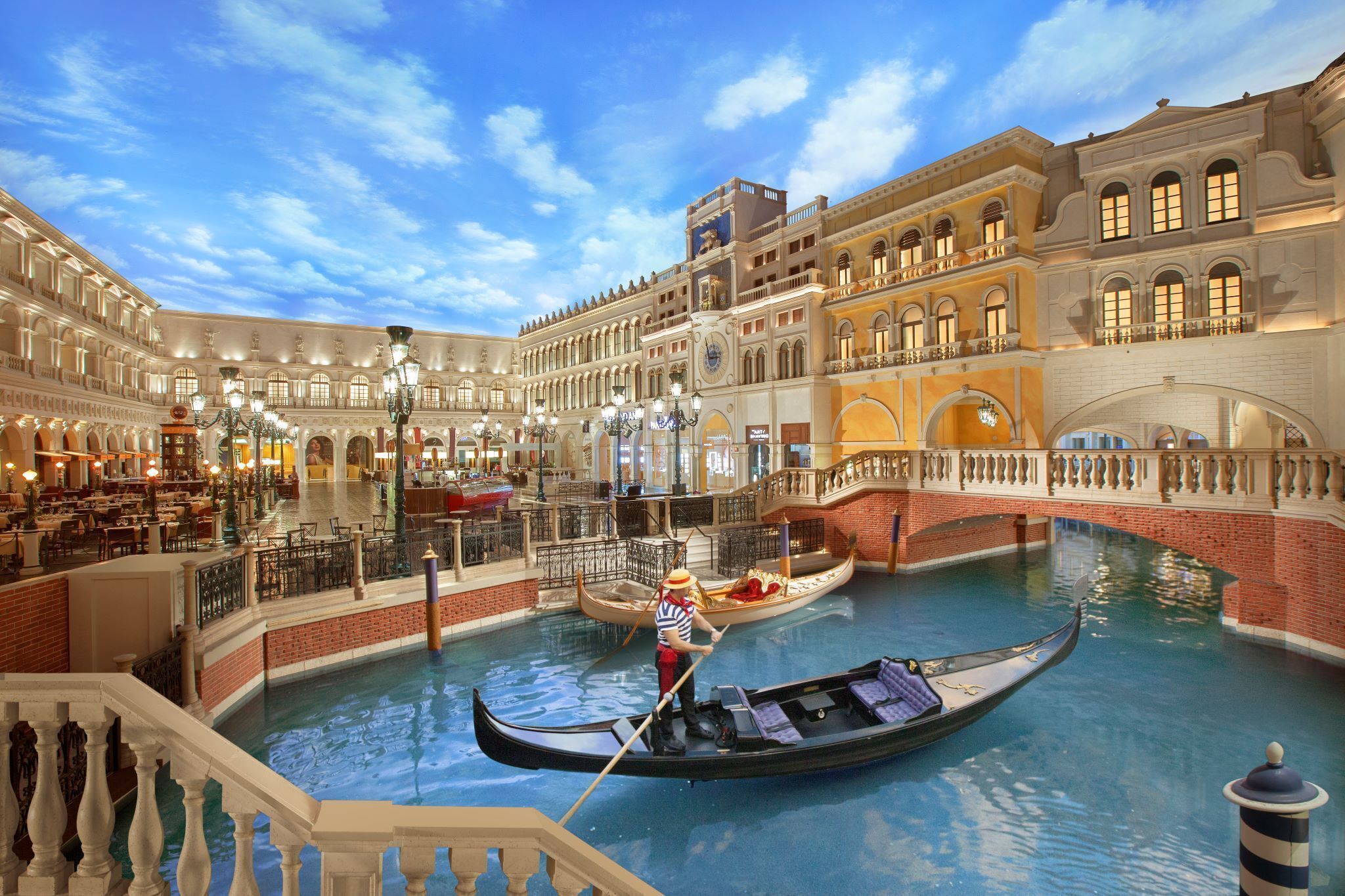 En indendørs kanal i venetiansk stil med gondoler og gondoliere, omgivet af prangende arkitektur og siddepladser.