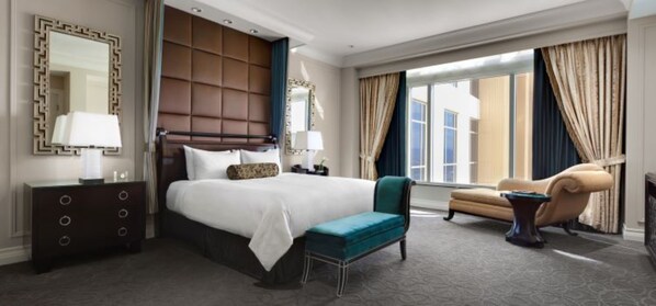 A hotel room with a large bed, a sitting area with a chair, a dresser, a mirror, and a window with curtains.