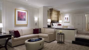 A modern hotel room with a beige sofa, a round coffee table, and a small side table. There is a bed with a headboard and a nightstand with a lamp.