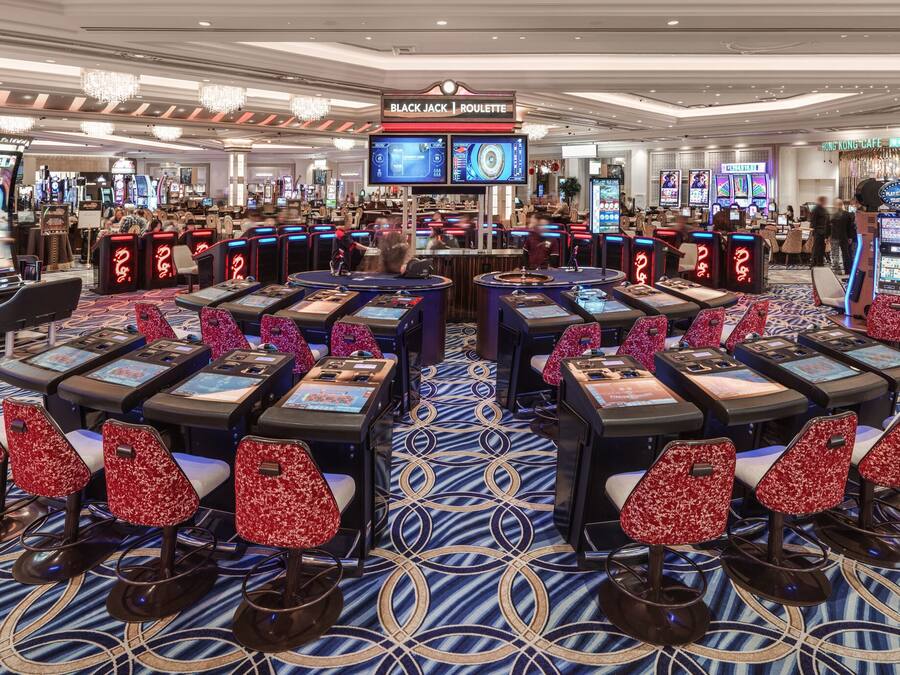 Une salle de casino avec de nombreuses tables de jeu et des chaises, un grand lustre et un panneau indiquant les jeux de «
Black Jack
» et de «
Roulette
».