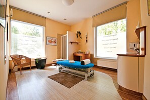 A treatment room with a massage table, a wicker chair, a small plant, and a window with a view of trees. - Łeba Hotel & Spa (Leba)