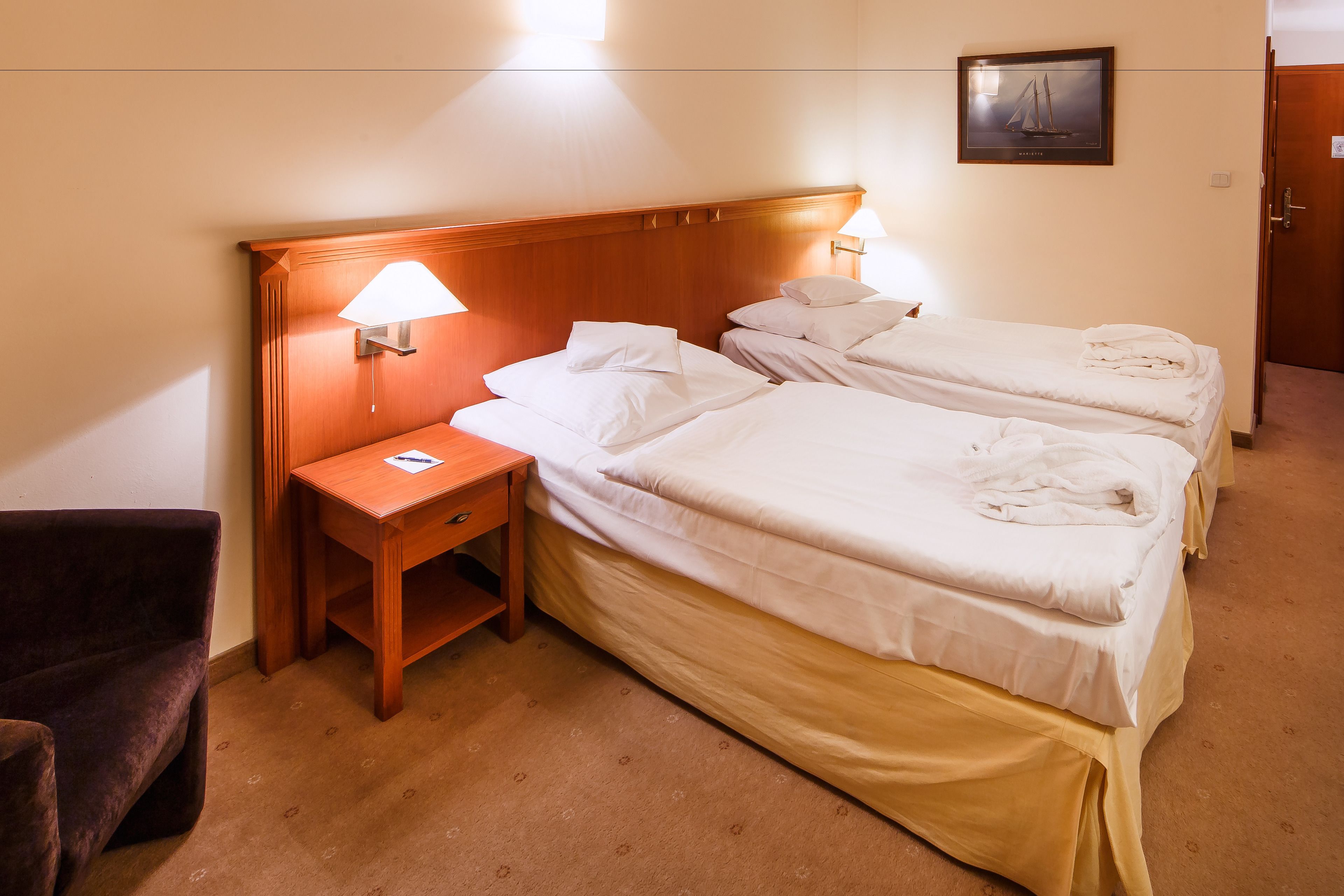 A hotel room with two beds, a nightstand, a lamp, a chair, and a framed picture on the wall.