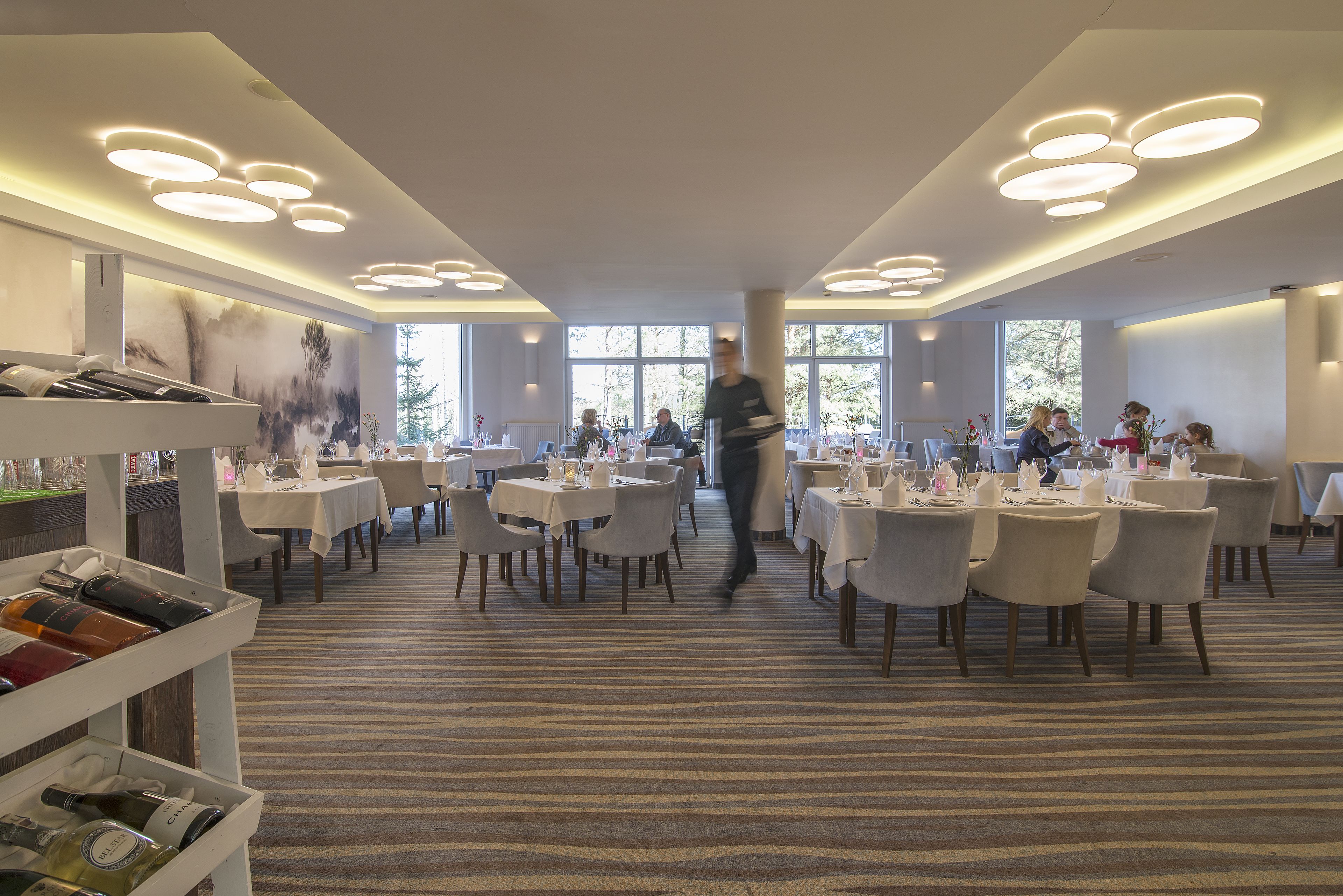 A spacious dining area with multiple tables and chairs, a waiter walking through, and a carpeted floor.