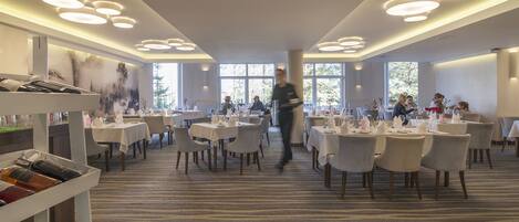 A spacious dining area with multiple tables and chairs, a waiter walking through, and a carpeted floor.