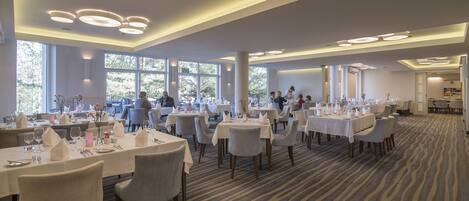 A spacious dining area with multiple round tables, each set with white tablecloths, plates, and cutlery. Large windows allow natural light to fill the room, and the carpet has a striped pattern.