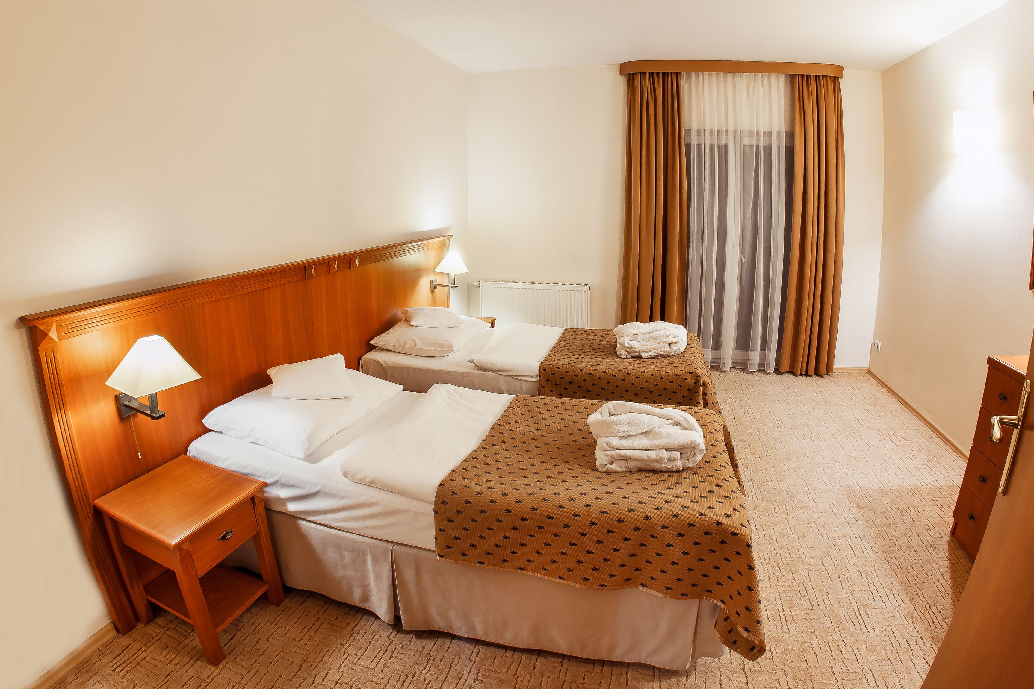 Habitación de hotel con dos camas, una mesita de noche, una lámpara, un radiador y un ventanal grande con cortinas.