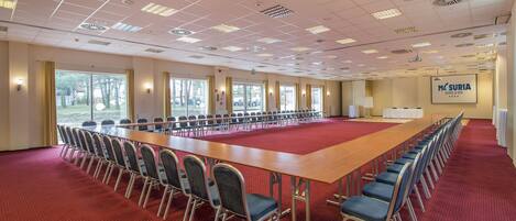 Un amplio salón de conferencias con una larga mesa y filas de sillas, ventanas grandes y un cartel que dice 'MÁSURIA HOTEL & SPA'.