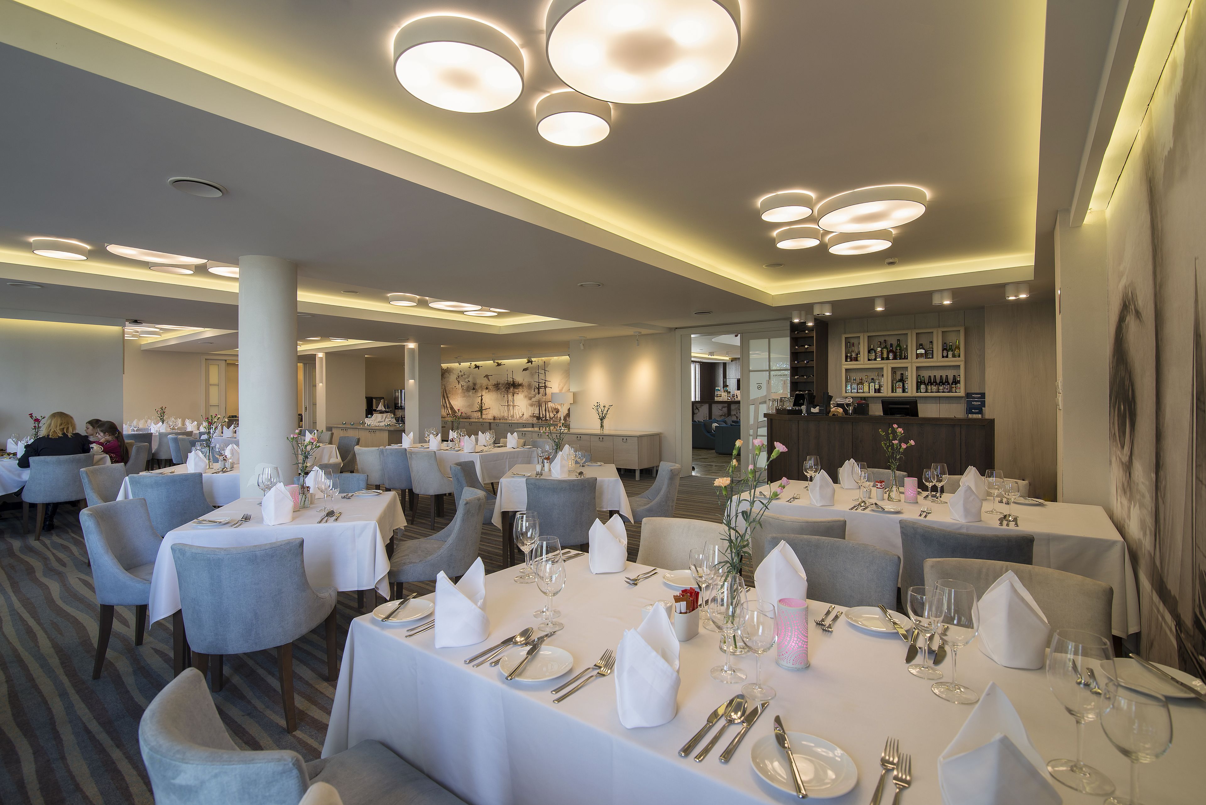 A well-lit dining area with round tables set for multiple guests, featuring white tablecloths and neatly arranged silverware.