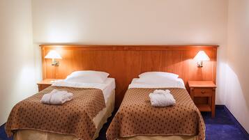 Habitación de hotel con dos camas, cada una con cabecero de madera, ropa blanca y colchas marrones estampadas. Hay dos mesitas de noche con lámparas y un pequeño cajón.